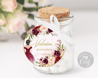 May include: A clear glass jar with a cork stopper, filled with white candies. A round tag is tied to the jar with a white ribbon, featuring a floral border, a graphic of a person in a dark red dress, and the text Valentina's Quinceañera 11.25.2021.