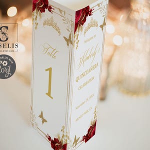 May include: A white table number card with gold accents, featuring the number 1. The card is decorated with gold butterflies, floral designs, and the text "Table" and "Kimberly's Quinceanera Celebration". Red floral accents adorn the corners.