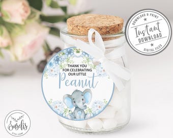 May include: A round paper gift tag featuring a watercolor illustration of a gray elephant, blue flowers, and green leaves. The text reads Thank You For Celebrating Our Little Peanut. The tag is tied with a white ribbon to a glass jar.