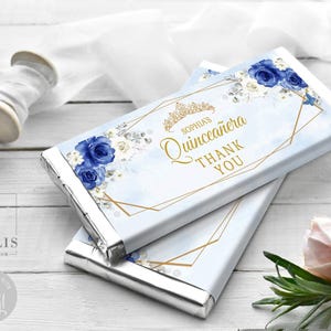May include: Two rectangular chocolate bars wrapped in custom paper featuring a light blue watercolor background, dark blue roses, white flowers, and gold geometric lines. The text reads Sophia's Quinceañera Thank You with a gold tiara icon.