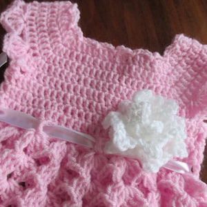 Puede incluir: Un vestido de beb&eacute; de ganchillo rosa con una flor de ganchillo blanca y una cinta de sat&eacute;n blanca.