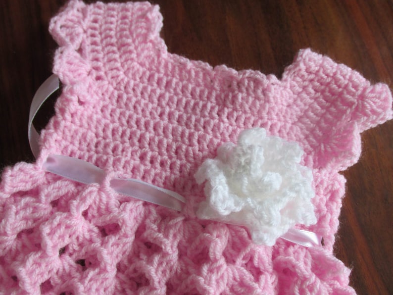 Puede incluir: Un vestido de beb&eacute; de ganchillo rosa con una flor de ganchillo blanca y una cinta de sat&eacute;n blanca.