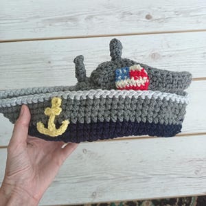May include: Handmade crochet boat in shades of gray and navy blue, featuring a yellow anchor and an American flag. The boat is a unique, handcrafted item, perfect for nautical-themed decor or as a child's toy.