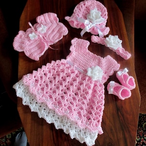 Puede incluir: Un vestido de beb&eacute; de ganchillo rosa con ribete de encaje blanco, gorro, c&aacute;rdigan, botines y diadema a juego de ganchillo rosa. El conjunto est&aacute; decorado con flores de ganchillo blancas.