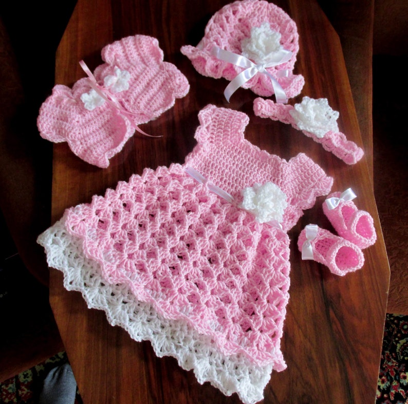 Puede incluir: Un vestido de beb&eacute; de ganchillo rosa con ribete de encaje blanco, gorro, c&aacute;rdigan, botines y diadema a juego de ganchillo rosa. El conjunto est&aacute; decorado con flores de ganchillo blancas.