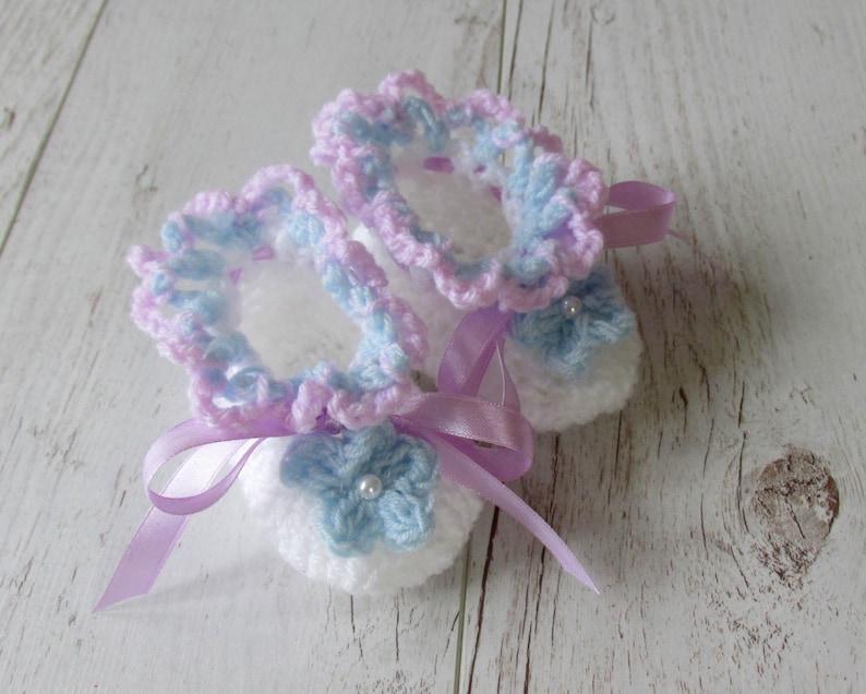 May include: A pair of crocheted baby booties in white, pink, and blue with a satin ribbon tied around them.