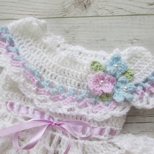 May include: White crocheted baby dress with a ruffled collar and delicate floral embellishments. The dress features a pink ribbon tied at the waist.