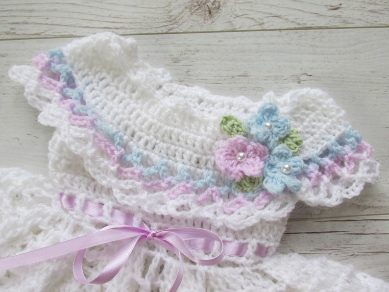 May include: White crocheted baby dress with a ruffled collar and delicate floral embellishments. The dress features a pink ribbon tied at the waist.