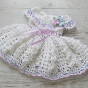 May include: White crocheted baby dress with a pink satin ribbon and a flower embellishment in shades of blue, pink, and green.