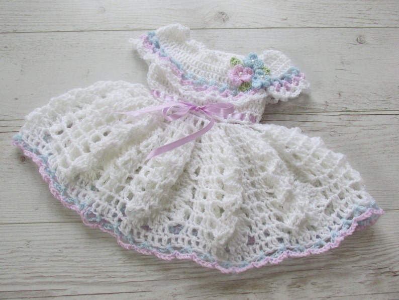 May include: White crocheted baby dress with a pink satin ribbon and a flower embellishment in shades of blue, pink, and green.