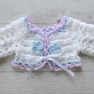 May include: White crocheted baby cardigan with a light blue and pink trim. The cardigan has a bow tie closure and two light blue crocheted flowers.
