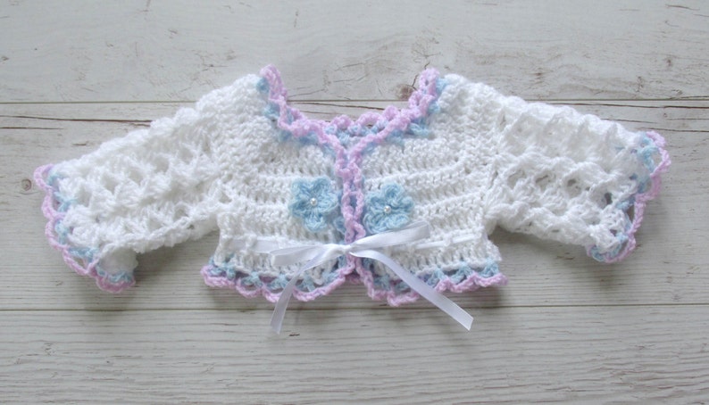 May include: White crocheted baby cardigan with a light blue and pink trim. The cardigan has a bow tie closure and two light blue crocheted flowers.