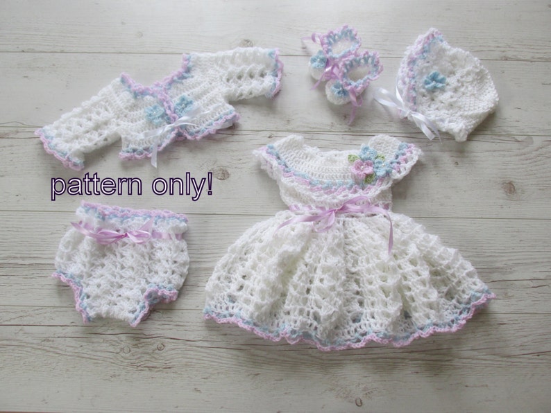 May include: A white crocheted baby outfit with a dress, cardigan, diaper cover, hat, and booties. The outfit is decorated with pink and blue accents and flowers.