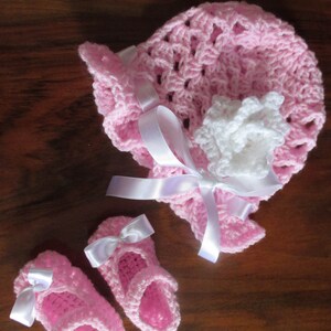 Puede incluir: Un gorro de beb&eacute; de ganchillo rosa con una flor de ganchillo blanca y una cinta de sat&eacute;n blanca. Dos zapatos de beb&eacute; de ganchillo rosa con lazos de sat&eacute;n blancos.