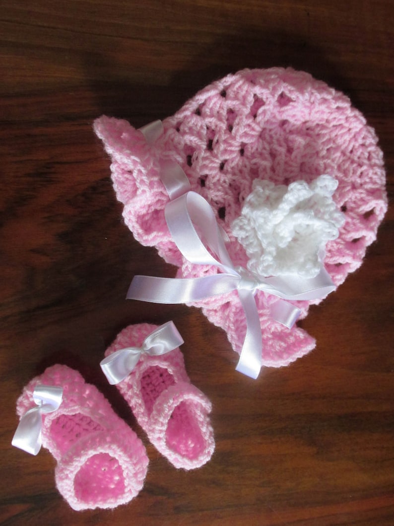 Puede incluir: Un gorro de beb&eacute; de ganchillo rosa con una flor de ganchillo blanca y una cinta de sat&eacute;n blanca. Dos zapatos de beb&eacute; de ganchillo rosa con lazos de sat&eacute;n blancos.
