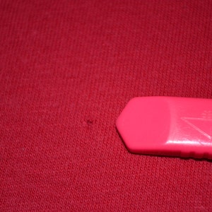 May include: A close-up of a red fabric surface with a small hole. A pink plastic utility knife is lying on the surface.