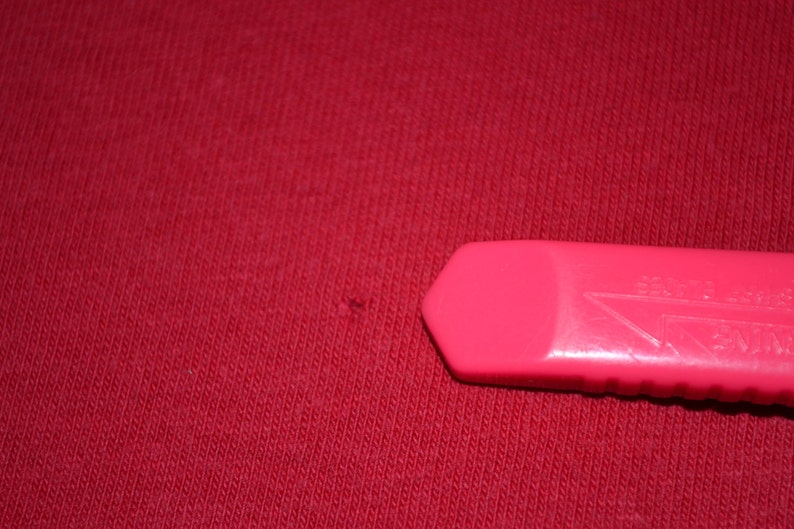 May include: A close-up of a red fabric surface with a small hole. A pink plastic utility knife is lying on the surface.