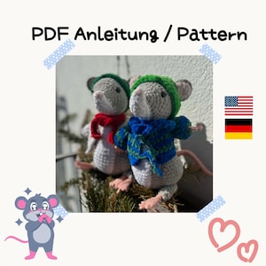 May include: Two crocheted gray mice wearing winter hats and scarves. The mouse on the left is wearing a red scarf and a brown hat. The mouse on the right is wearing a blue scarf and a green hat. The mice are sitting on a bed of green pine needles.