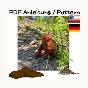 Könnte beinhalten: Eine gehäkelte braune Kreatur mit dunkelroten Armen und Beinen sitzt im Gras. Das Bild enthält den Text "PDF Anleitung / Pattern" und die amerikanische Flagge. Die Kreatur ist von Gras und Erde umgeben.