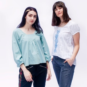 May include: Two women wearing different styles of blouses. The woman on the left is wearing a light blue blouse with long sleeves and a square neckline. The woman on the right is wearing a white blouse with short sleeves and a blue embroidered design.