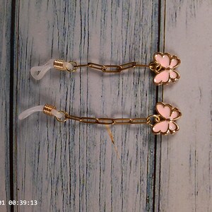 May include: A pair of gold-toned eyeglass chains featuring light pink enamel butterfly charms at the ends. Each chain includes a gold-colored metal link segment and a clear, flexible silicone loop attachment designed to secure onto eyewear frames.
