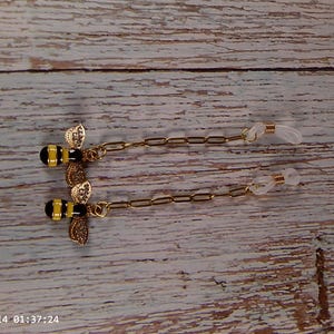 May include: A pair of gold-toned eyeglass chains featuring bee charms with yellow and black striped bodies and clear crystal-encrusted wings. Each chain connects to a clear silicone loop designed to secure onto the temples of eyeglasses.