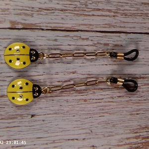 May include: A pair of eyeglass chain charms featuring yellow ladybug pendants with black spots and black heads. Each charm is attached to a gold-toned metal chain link that connects to a black rubber loop with a gold-toned metal coil accent.