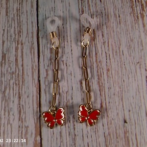 May include: Two eyeglass chains with gold-colored links and red butterfly charms. The chains have clear rubber loops for attaching to glasses. The background is a light-colored wooden surface.