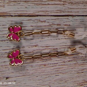 May include: Gold-tone eyeglass chain with pink butterfly charms. The chain features a delicate link design and clear rubber loops for attaching to glasses. The butterfly charms have a gold outline and are filled with a vibrant pink color.