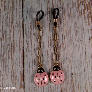 May include: Two eyeglass chains with black loops, gold-colored chains, and pink ladybug charms with black spots. The chains hang against a light-colored, wood-grain background.