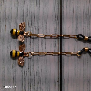 May include: A pair of gold-toned eyeglass chains featuring bee charms with black and yellow striped bodies and clear rhinestone-encrusted wings. Each chain includes a gold-colored metal link design and black rubber loops at the ends for attachment.