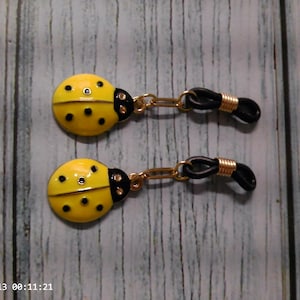 May include: A pair of eyeglass chain connectors featuring yellow ladybug charms with black spots and black heads. Each charm is attached to a gold-toned metal link connected to a black rubber loop with a gold-toned metal coil for securing eyewear.