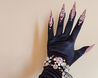 May include: A long black satin glove with a jeweled bracelet. The glove has a gathered texture and the fingers are extended with long, pointed, pink jeweled nails.
