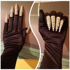 Puede incluir: Guantes de satén negro con uñas largas, puntiagudas y adornadas con joyas. Las uñas están cubiertas de piedras de imitación iridiscentes.