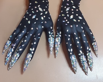 May include: A pair of black gloves adorned with numerous iridescent rhinestones scattered across the surface. The fingertips are embellished with long, pointed, crystal-like accents, creating a dramatic and glamorous effect.