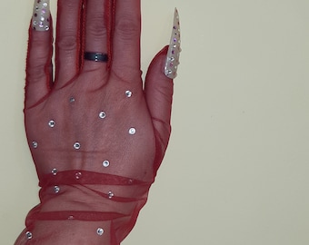 May include: A hand wearing a red mesh glove with small silver dots. The hand is wearing long, pointed, clear acrylic nails with silver and purple rhinestones.