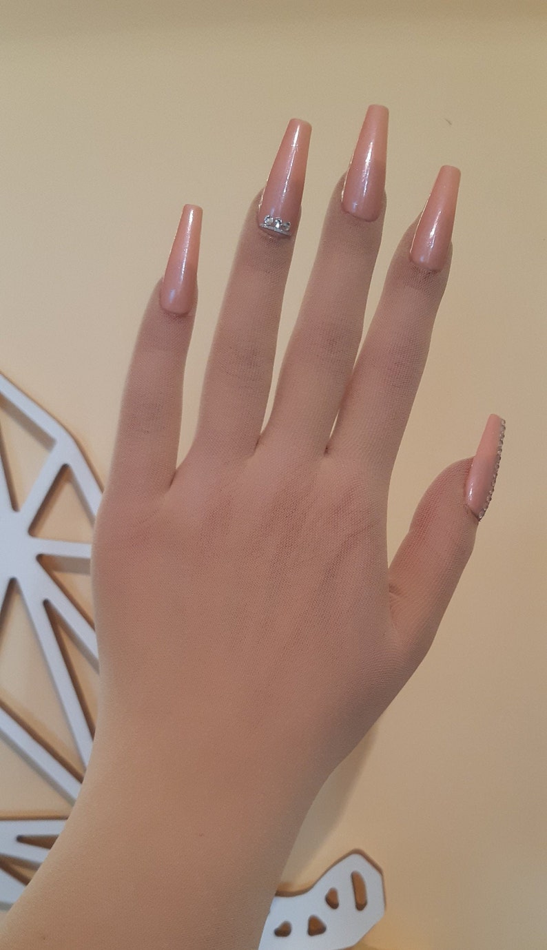 May include: A hand with long, nude-colored acrylic nails with rhinestones. The nails are long and pointed.