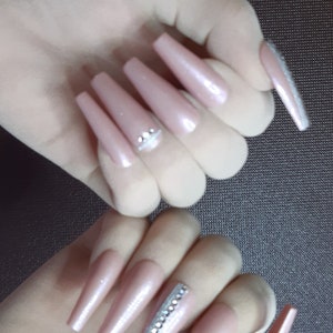 May include: Two hands with long, nude-colored, almond-shaped acrylic nails. The nails are decorated with rhinestones. One hand has a single row of rhinestones on one nail, while the other hand has a single row of rhinestones on two nails.