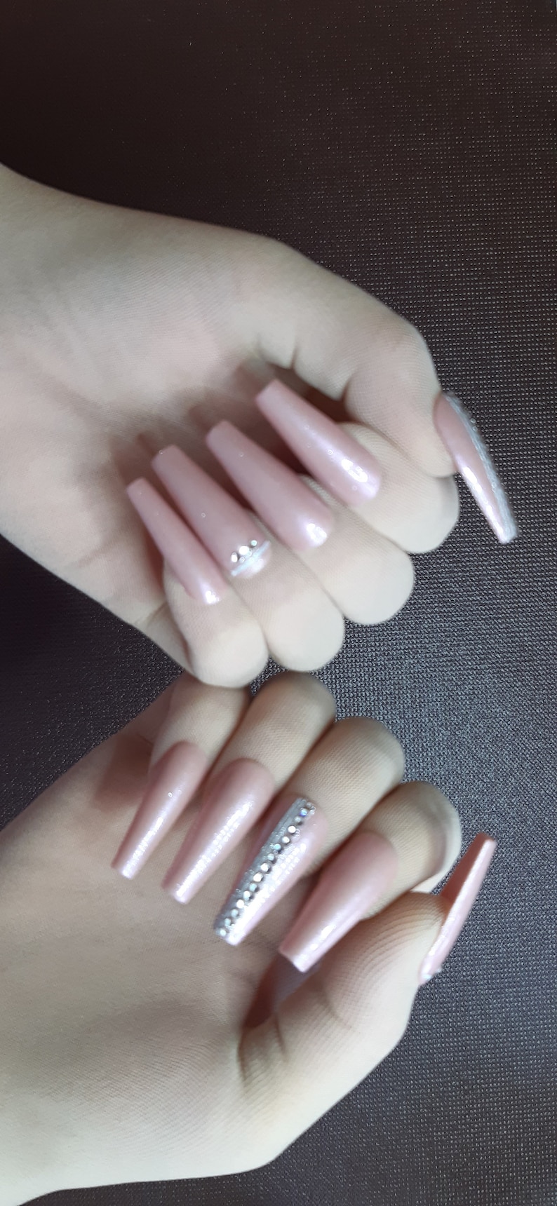 May include: Two hands with long, nude-colored, almond-shaped acrylic nails. The nails are decorated with rhinestones. One hand has a single row of rhinestones on one nail, while the other hand has a single row of rhinestones on two nails.