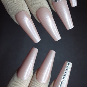 May include: A set of long, nude-colored, artificial nails with a shimmery finish. Some nails have a row of small, clear rhinestones.