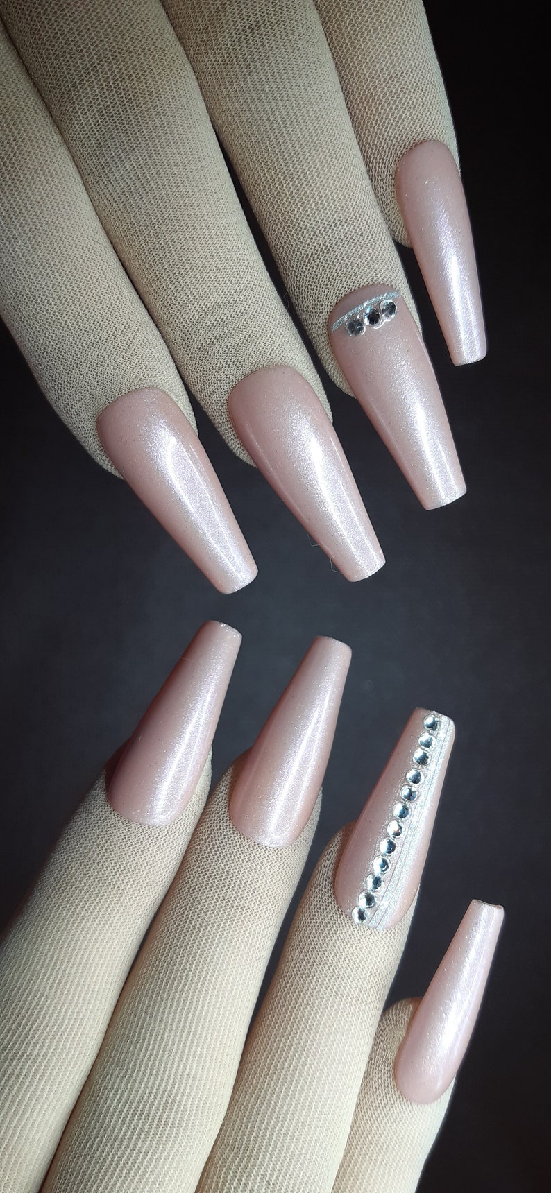 May include: A set of long, nude-colored, artificial nails with a shimmery finish. Some nails have a row of small, clear rhinestones.