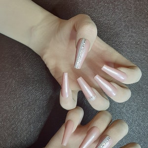 May include: Two hands wearing nude colored gloves with long, nude colored, almond shaped fake nails with rhinestones on the tips.