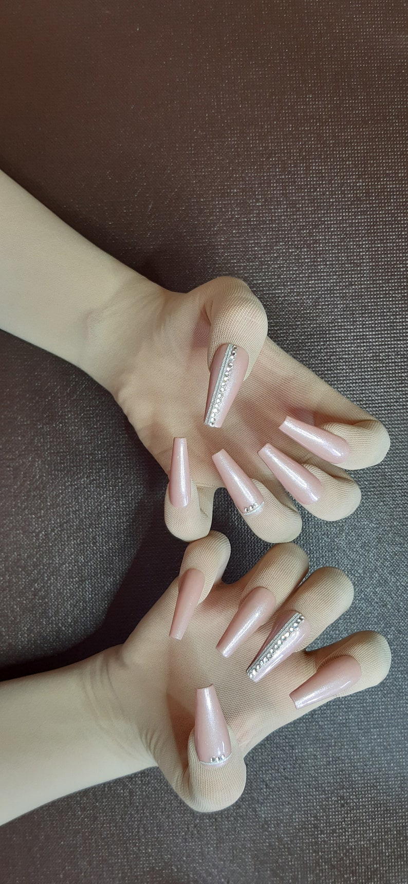 May include: Two hands wearing nude colored gloves with long, nude colored, almond shaped fake nails with rhinestones on the tips.