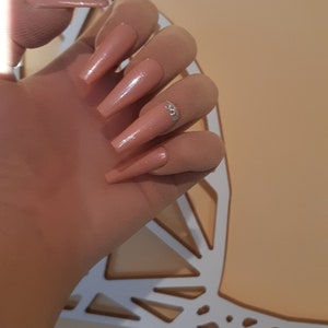 May include: Close-up of a hand with long, nude-colored, almond-shaped acrylic nails with a single rhinestone accent on one nail.
