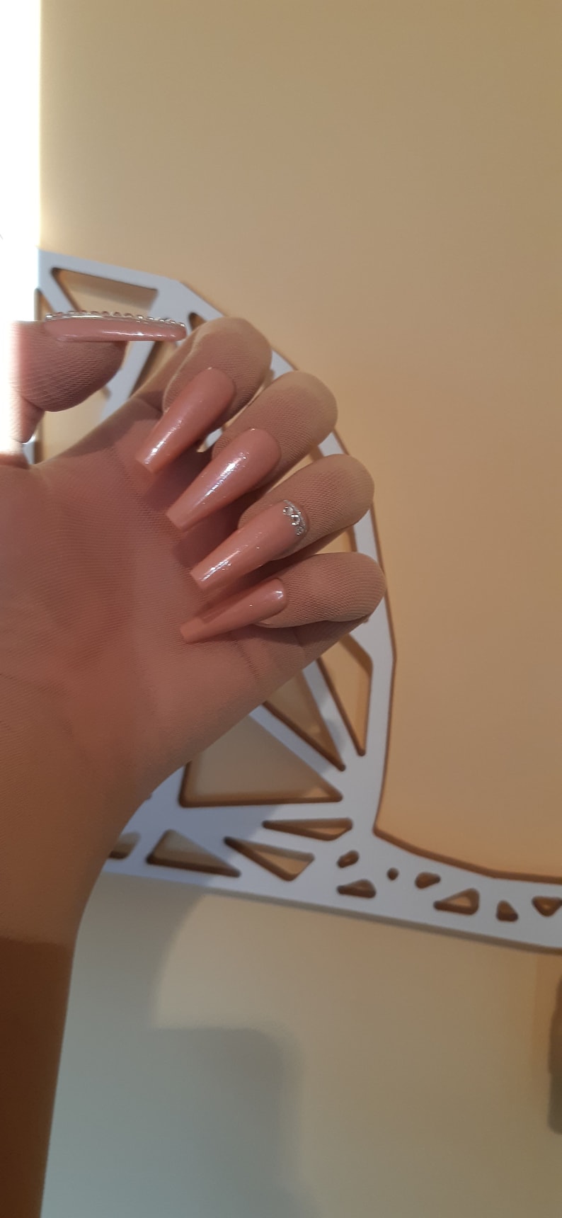 May include: Close-up of a hand with long, nude-colored, almond-shaped acrylic nails with a single rhinestone accent on one nail.