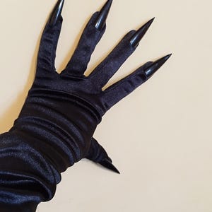 Puede incluir: Guantes de satén negro con uñas negras largas y puntiagudas. Los guantes son de color negro oscuro y sólido y tienen una textura suave y brillante. Las uñas son puntiagudas y se extienden más allá de las puntas de los dedos.