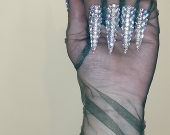 May include: A hand wearing a dark green sheer mesh glove with long, pointed, clear acrylic nails decorated with iridescent rhinestones.
