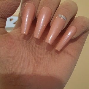 May include: A set of five long, nude-colored fake nails with a clear glitter accent on one nail.