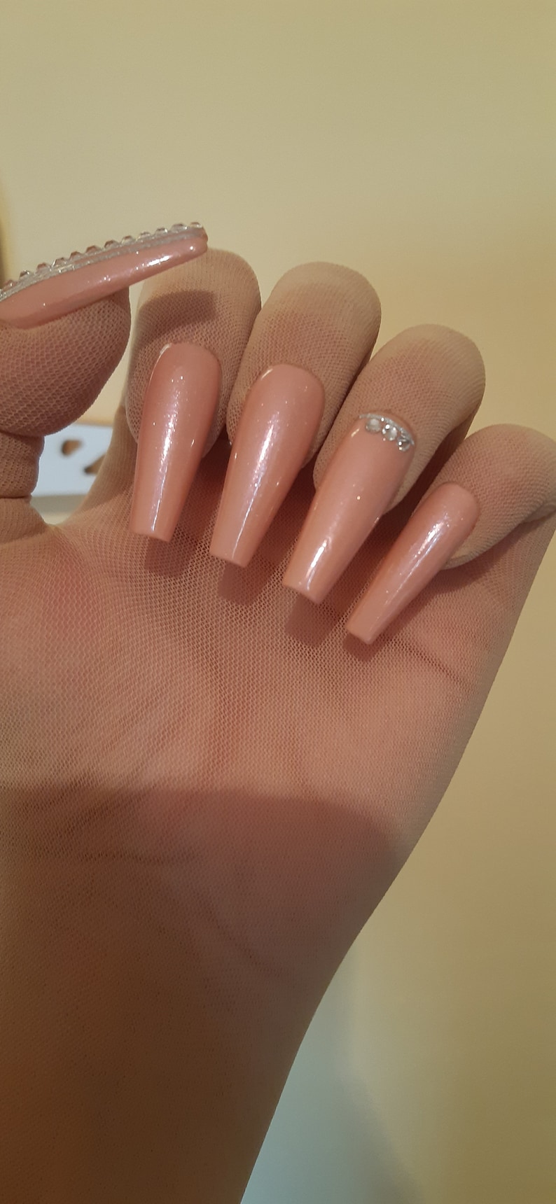 May include: A set of five long, nude-colored fake nails with a clear glitter accent on one nail.