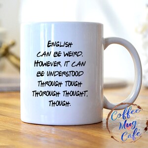 May include: White ceramic coffee mug with black text that reads "English can be weird. However, it can be understood through tough thorough thought, though."